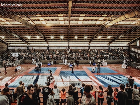 A large indoor arena with a high ceiling, filled with an audience seated on bleachers. In the center, two fighters are engaged in a martial arts competition on blue and red mats. The scene is captured through a mesh fence, adding a textured foreground.