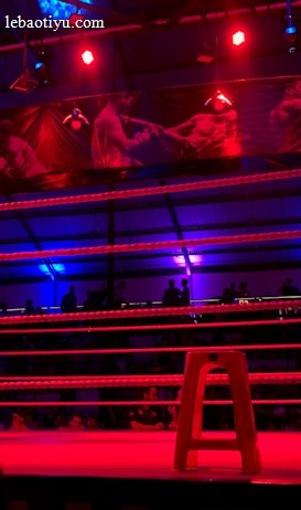 An empty boxing ring is illuminated by dramatic red and blue spotlights inside a dimly-lit arena. A small stool is placed in the center of the ring, surrounded by ropes. The arena is adorned with banners depicting action scenes, and a few silhouettes of spectators can be seen in the background.